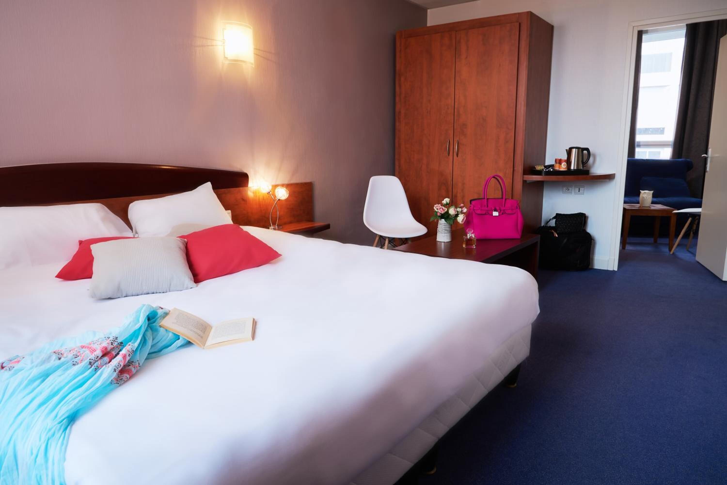 A hotel room with a large bed, a wardrobe, a desk with a chair, and a small table with a kettle.