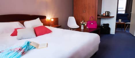 A hotel room with a large bed, a wardrobe, a desk with a chair, and a small table with a kettle.
