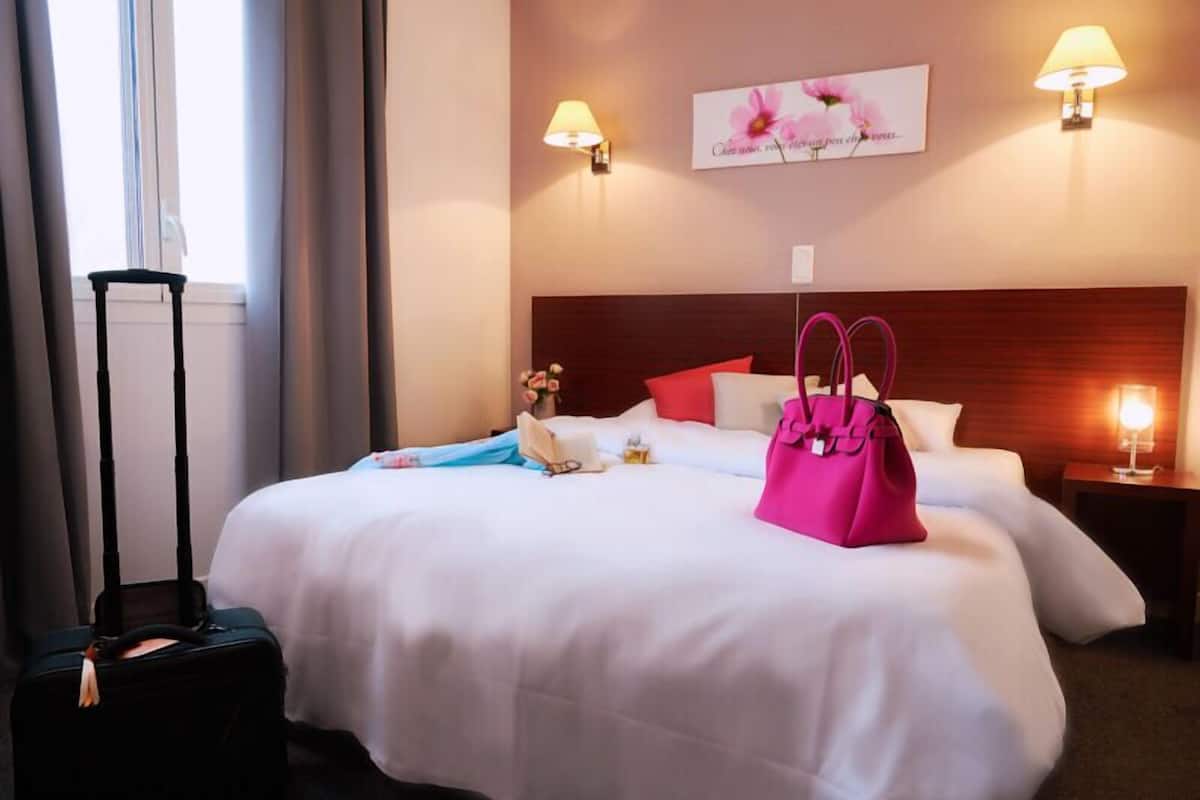 Habitación de hotel con una cama grande, dos lámparas de noche, una mesita de noche, una maleta y una ventana con cortinas.