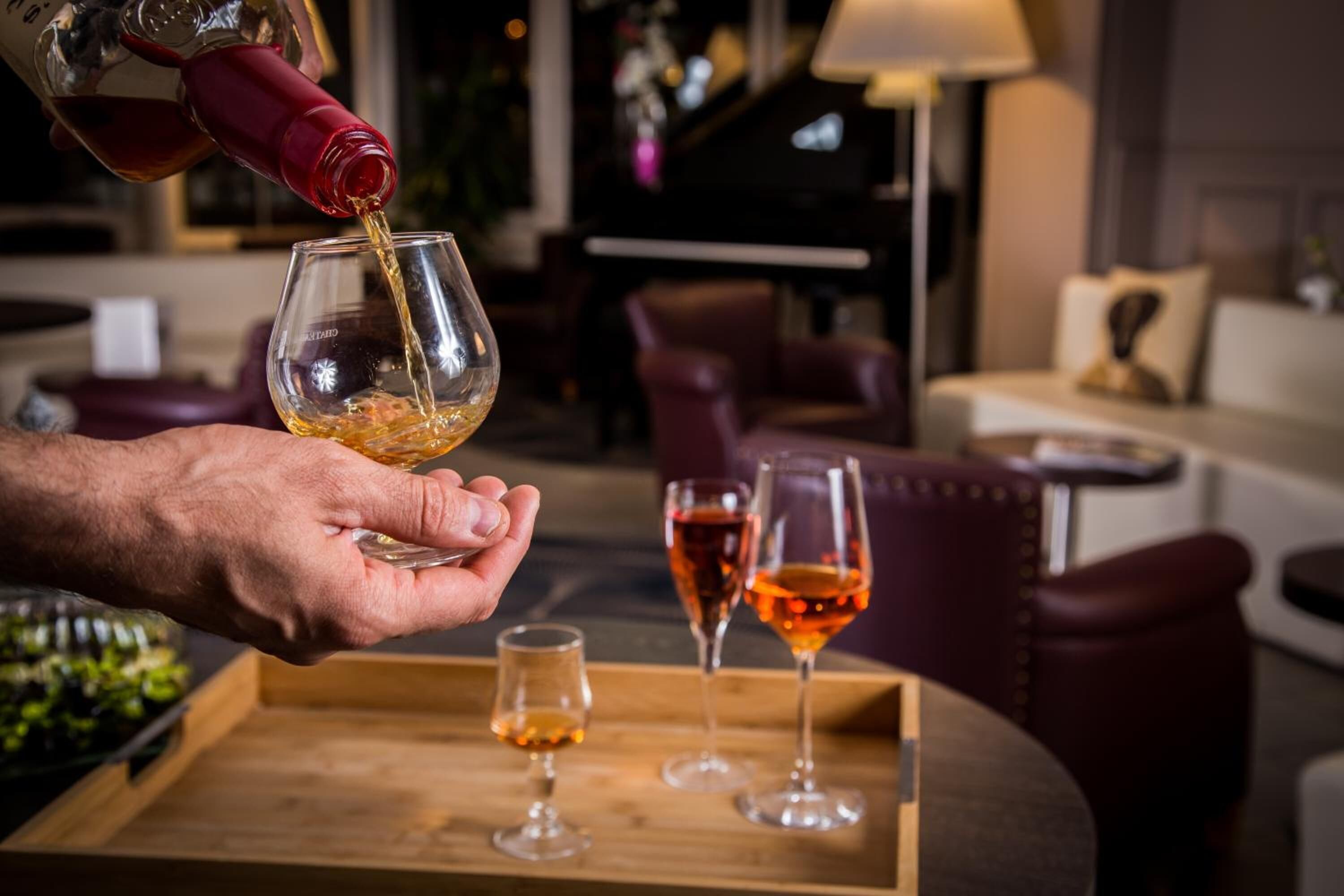 A person pouring a drink into a glass, with other glasses and a tray in the background.