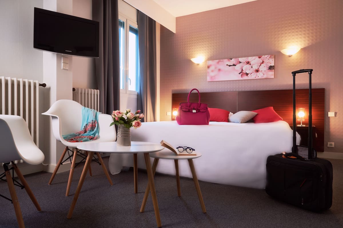 Habitación de hotel con cama, televisor, mesa con un jarrón de flores, una silla y una maleta.