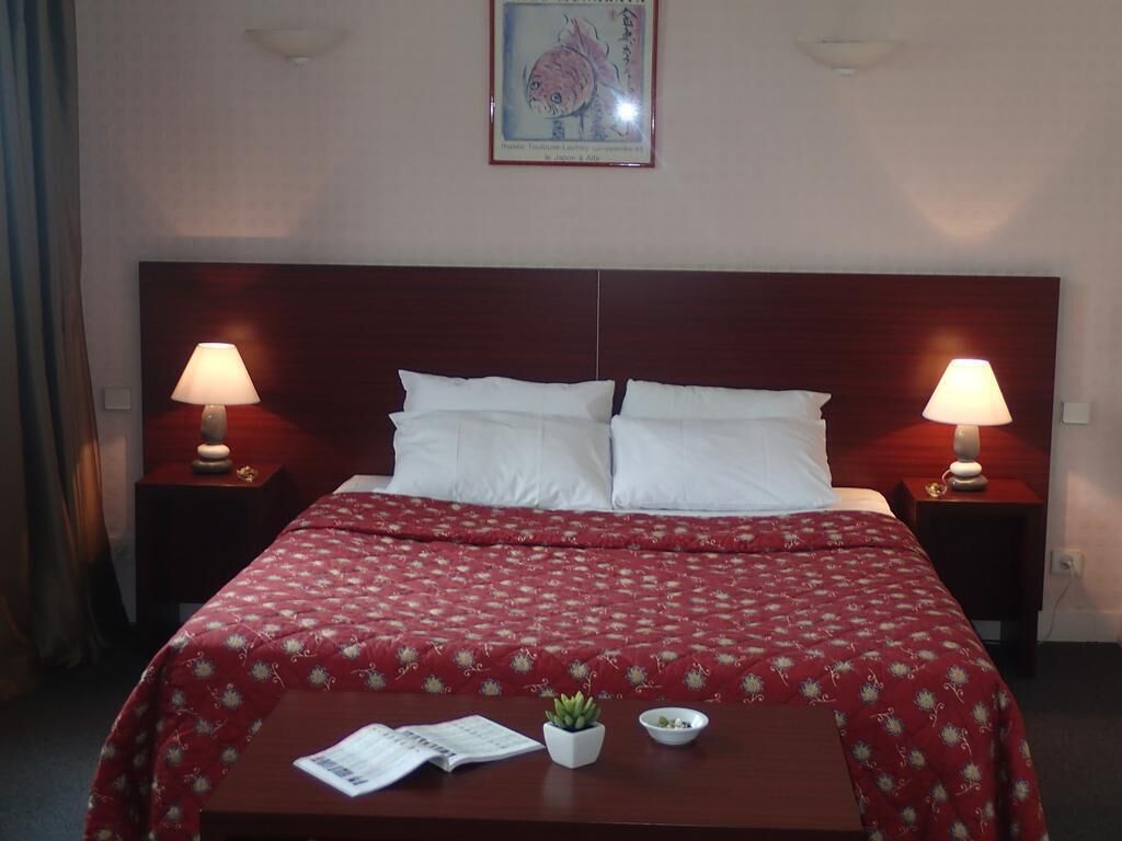 Une chambre d’hôtel avec un lit double, deux tables de chevet avec des lampes, une petite table avec une plante et un bol, et un tableau encadré au mur.