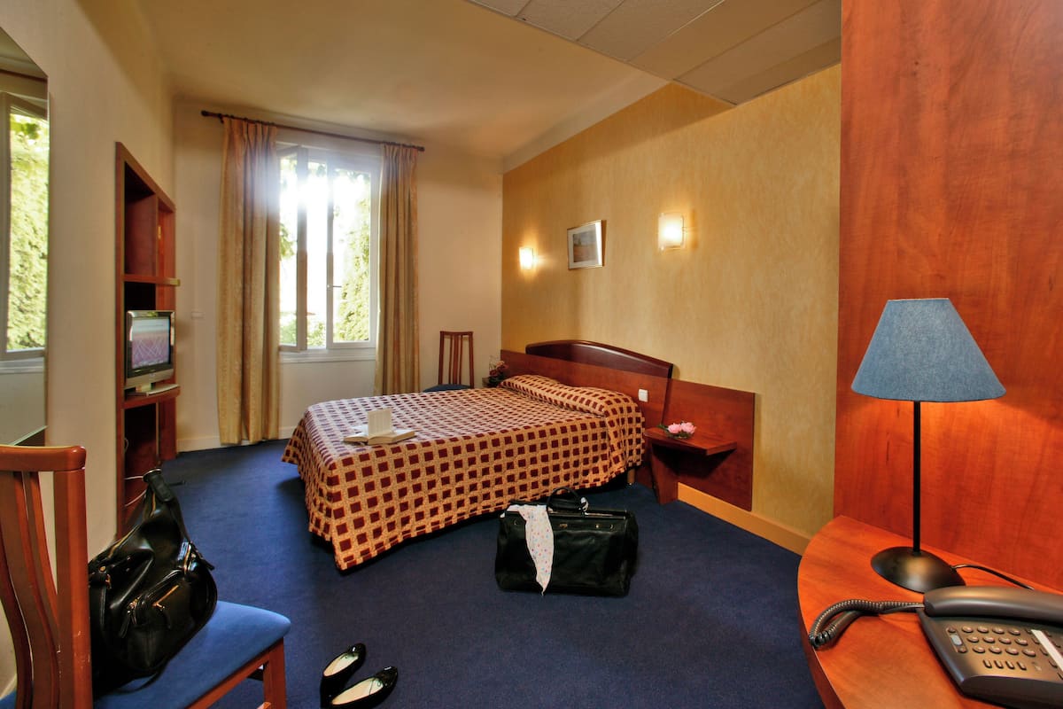Una habitación de hotel con una cama, un escritorio con lámpara, un teléfono, una silla, una maleta y un televisor sobre un mueble.
