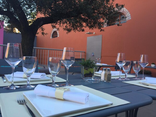 Una mesa preparada para una comida, con copas de vino, platos y cubiertos, en un espacio al aire libre con un árbol y un edificio al fondo.