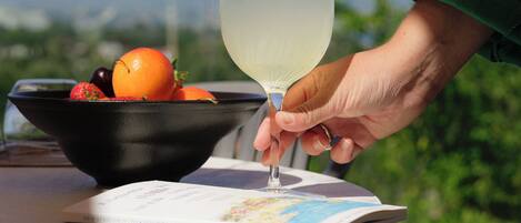 A person holding a glass of a clear liquid with a lime slice, a bowl of fruit, and a book on a table with a scenic view in the background.