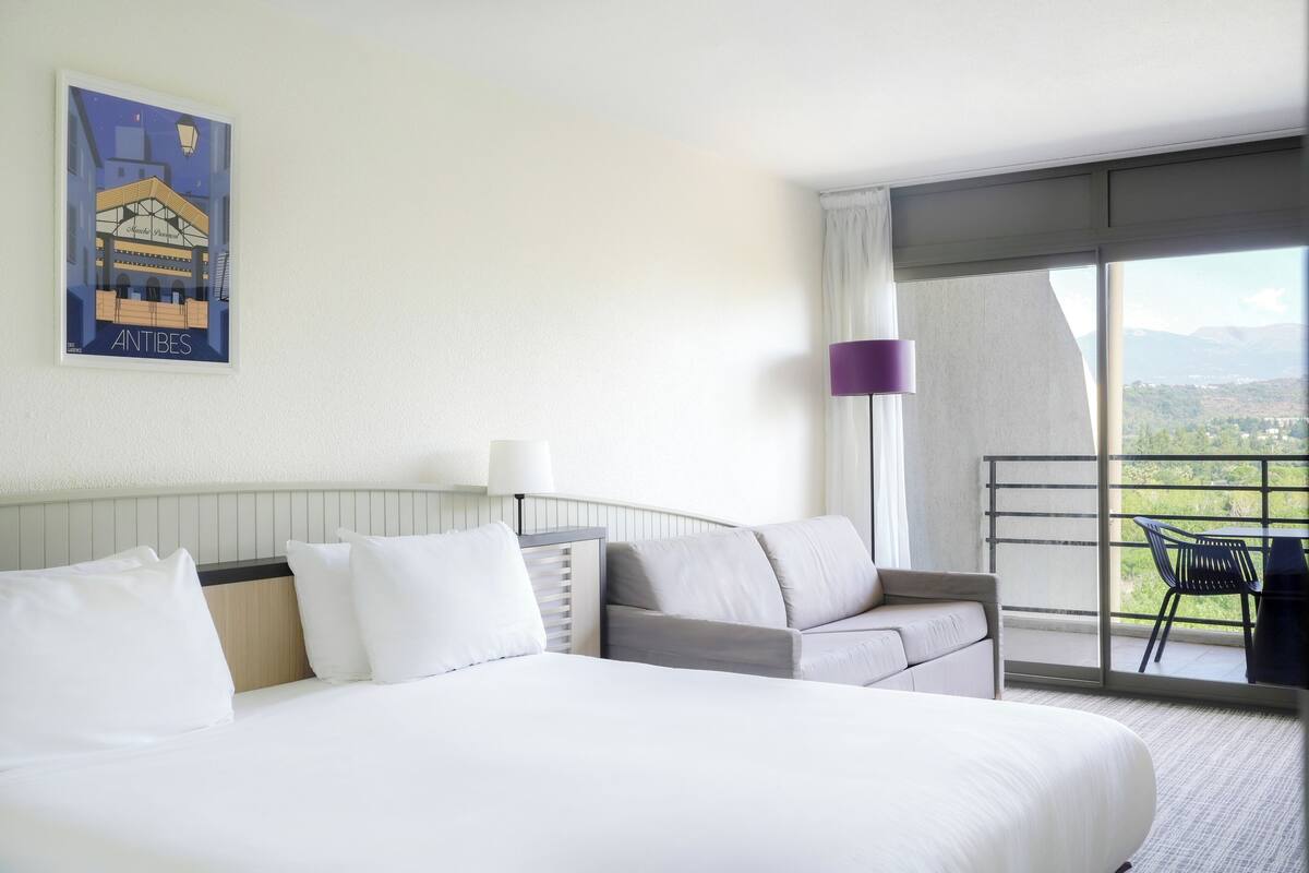 A hotel room with a large bed, a sofa, a balcony with a view, and a painting on the wall.