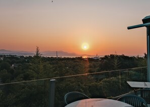 Vista al atardecer desde un balcón con mesa y sillas, con un paisaje de árboles y montañas a lo lejos.