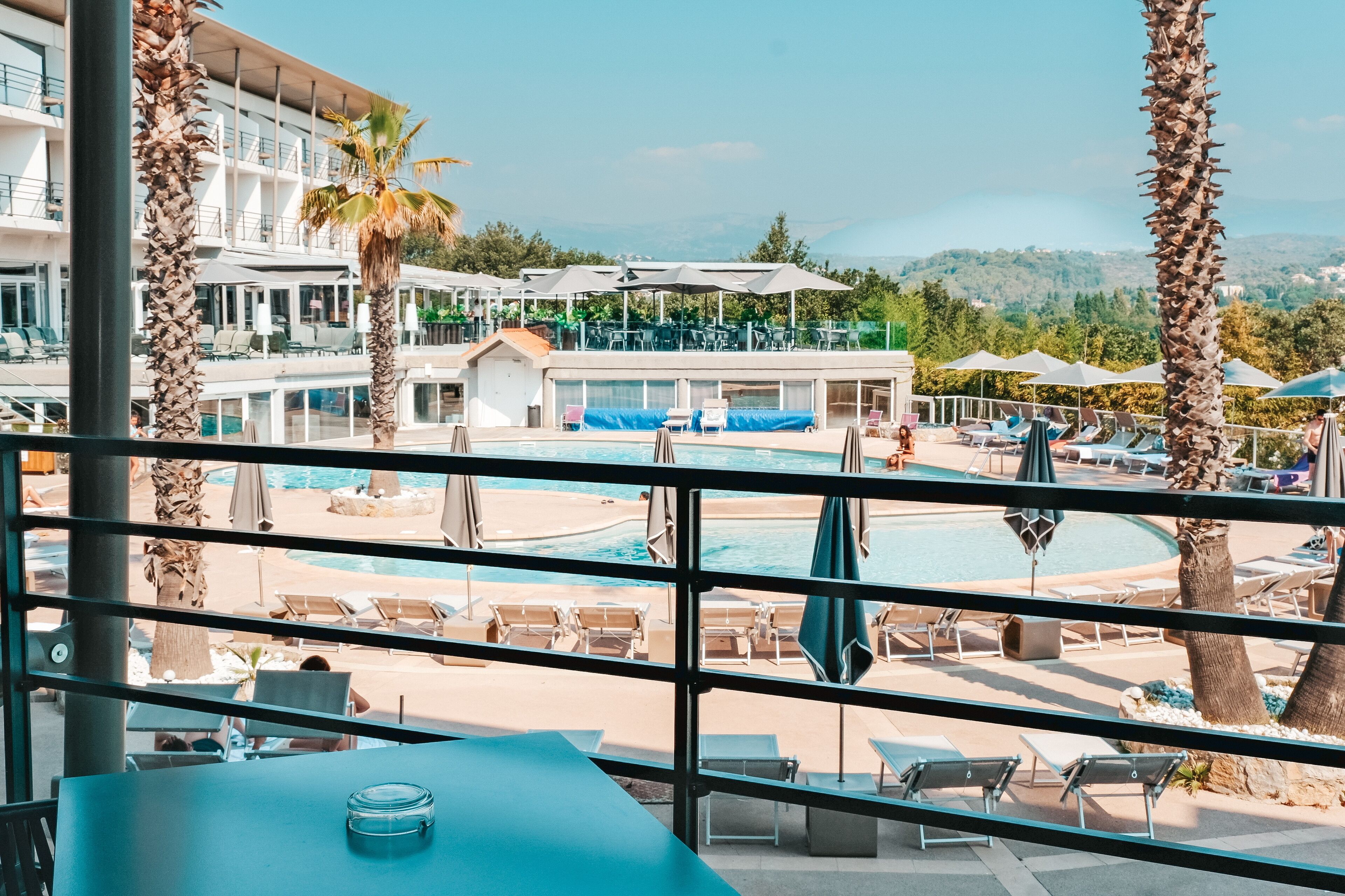 Un espace piscine avec des chaises longues et des parasols, une vue sur la piscine et un bâtiment avec des balcons en arrière-plan.