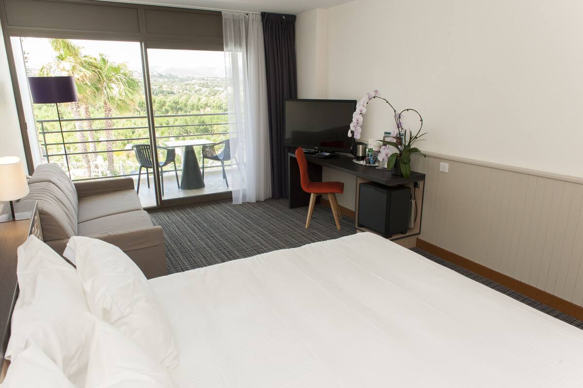 A hotel room with a bed, a desk, a chair, a TV, and a balcony with a view of greenery.