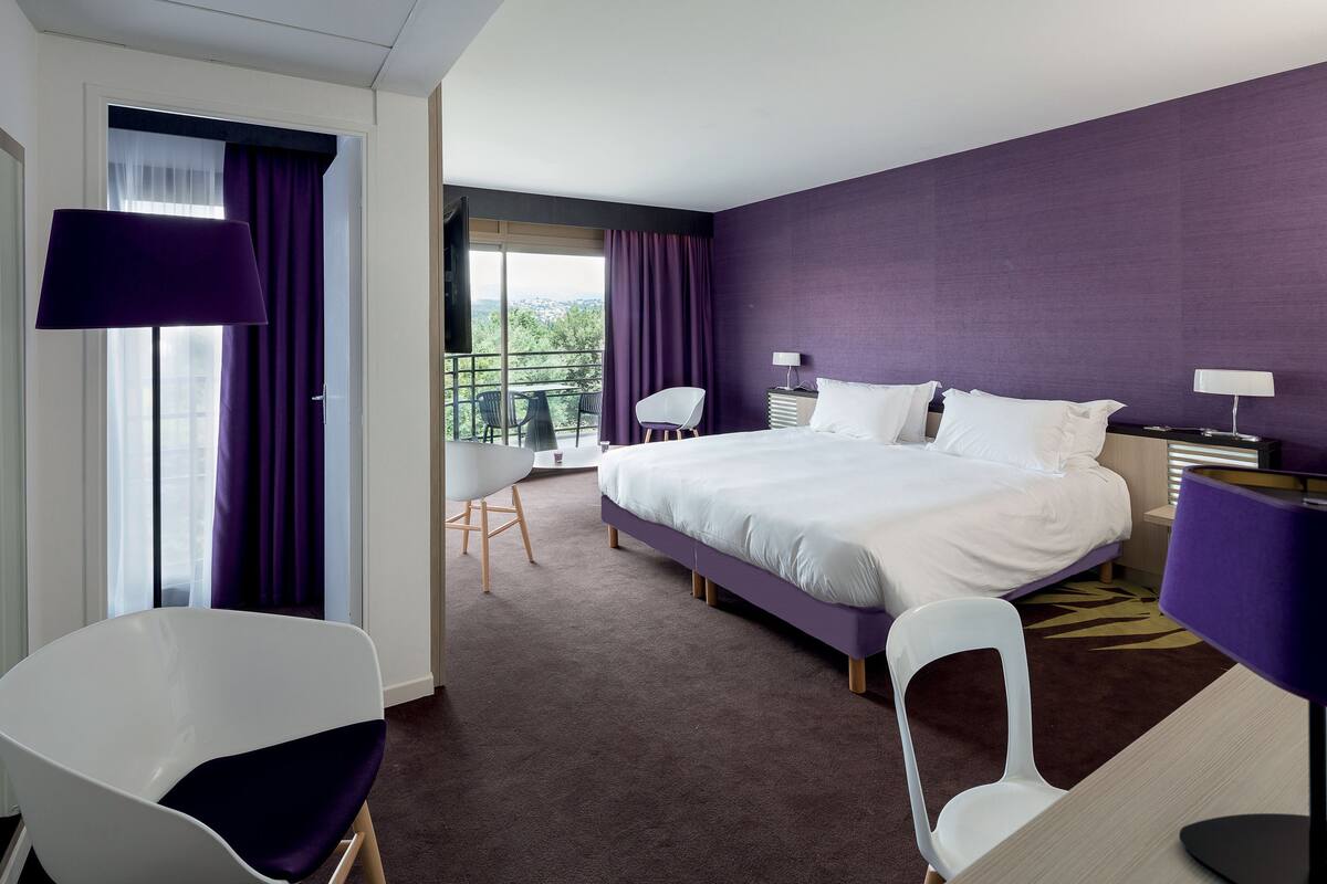 A hotel room with a large bed, two chairs, a balcony with a view, and purple walls.