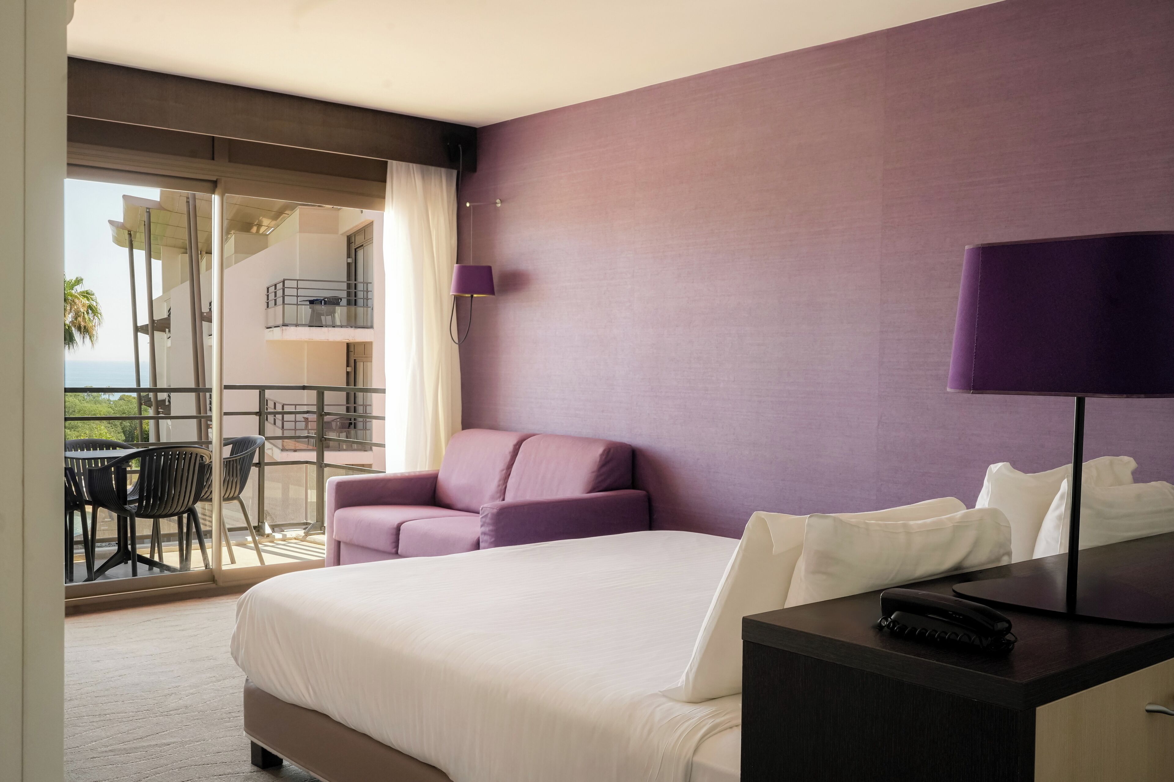 Habitación de hotel con una cama, un escritorio con teléfono, una silla morada y un balcón con mesa y sillas.