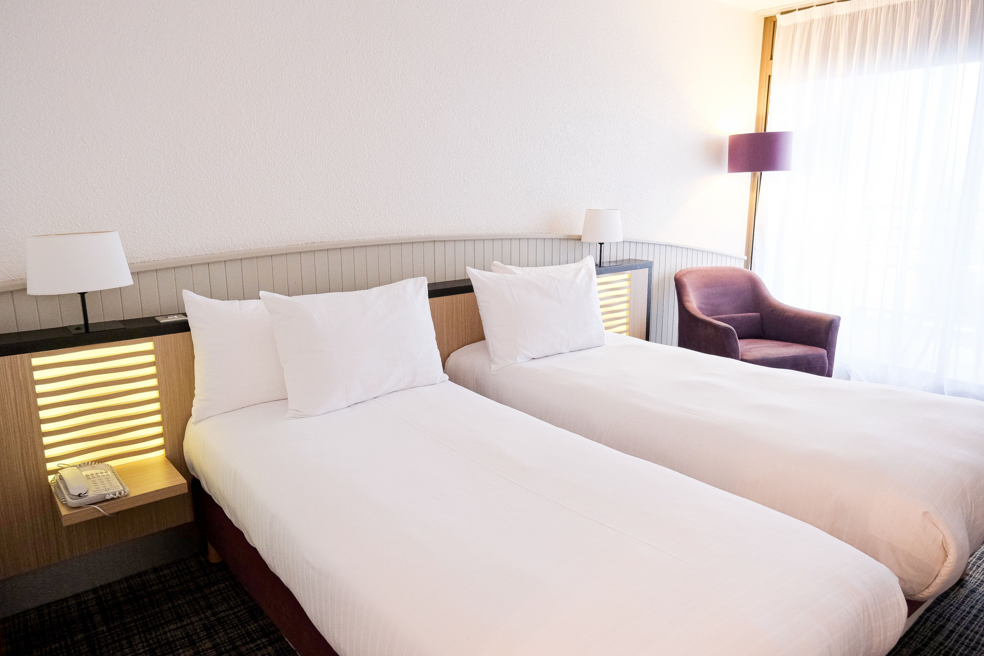 Een hotelkamer met twee bedden, bedlampen, een stoel en een telefoon op een nachtkastje.