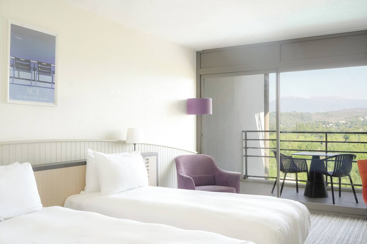 A hotel room with two beds, a purple armchair, a table with chairs, a balcony with a view of the countryside, and a poster on the wall.