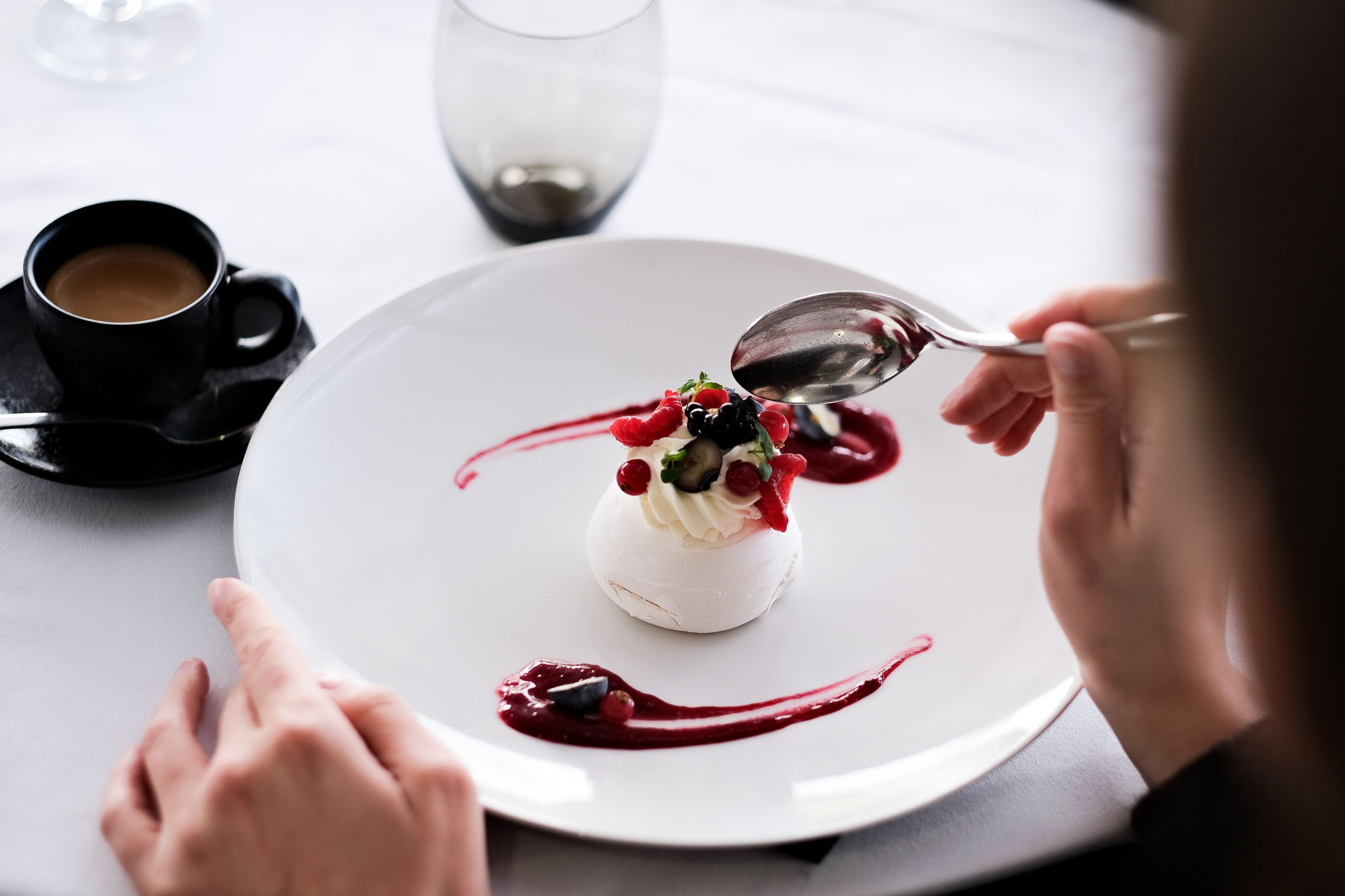 A dessert on a white plate with a spoon, a cup of coffee, and a glass on a table.
