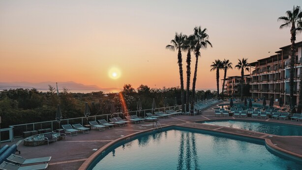 Zona de piscina en un resort con sillones de descanso y palmeras, rodeada por un edificio con varios balcones.