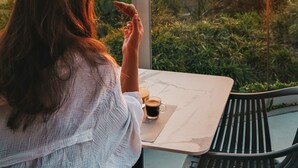 Una persona sentada a una mesa con una bebida, contemplando un paisaje pintoresco al atardecer.