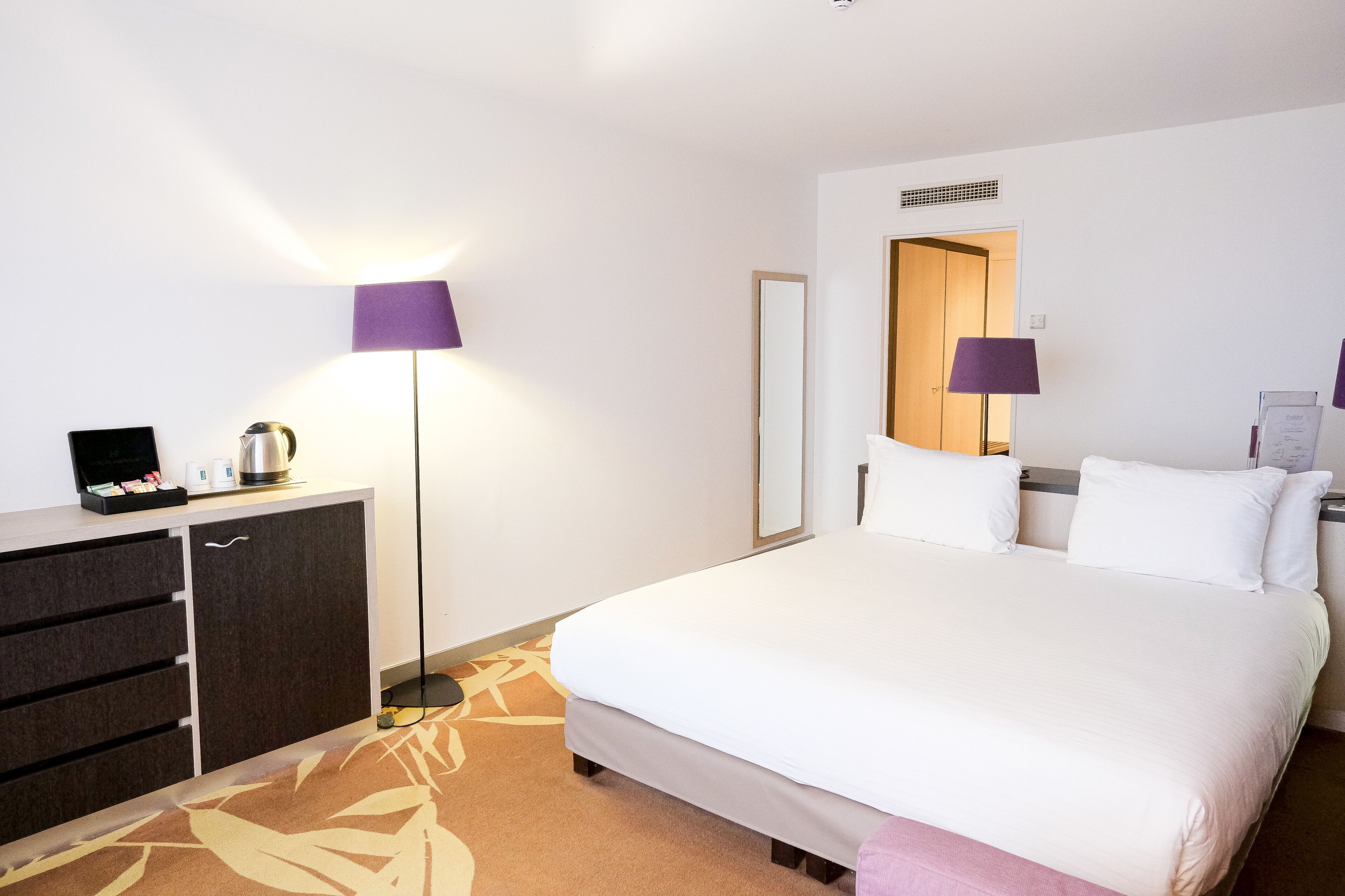 A hotel room with a large bed, a bedside table, a dresser, and two lamps.