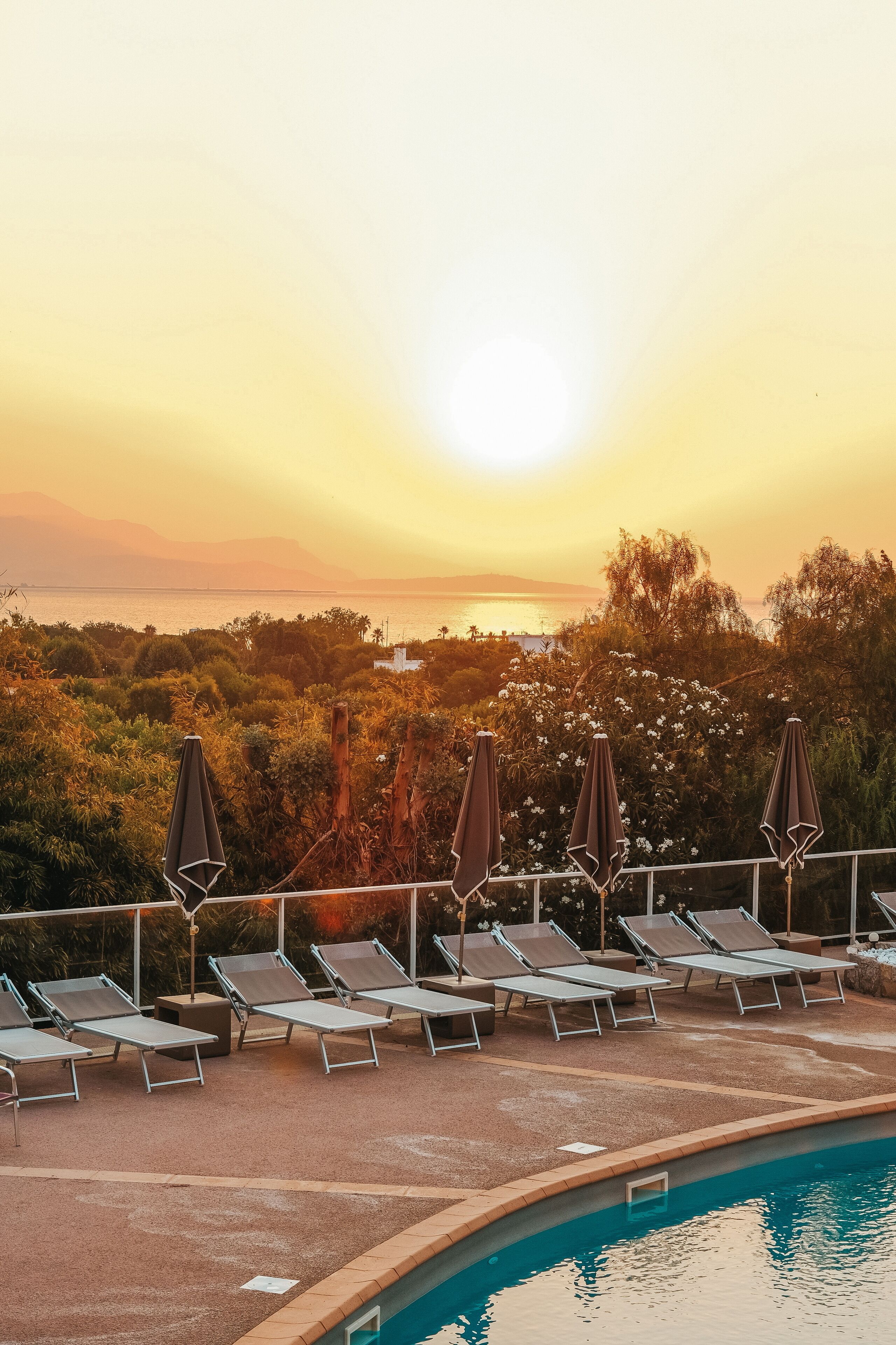 Área de piscina com espreguiçadeiras e guarda-sóis, com vista para um corpo de água e colinas durante o pôr do sol.