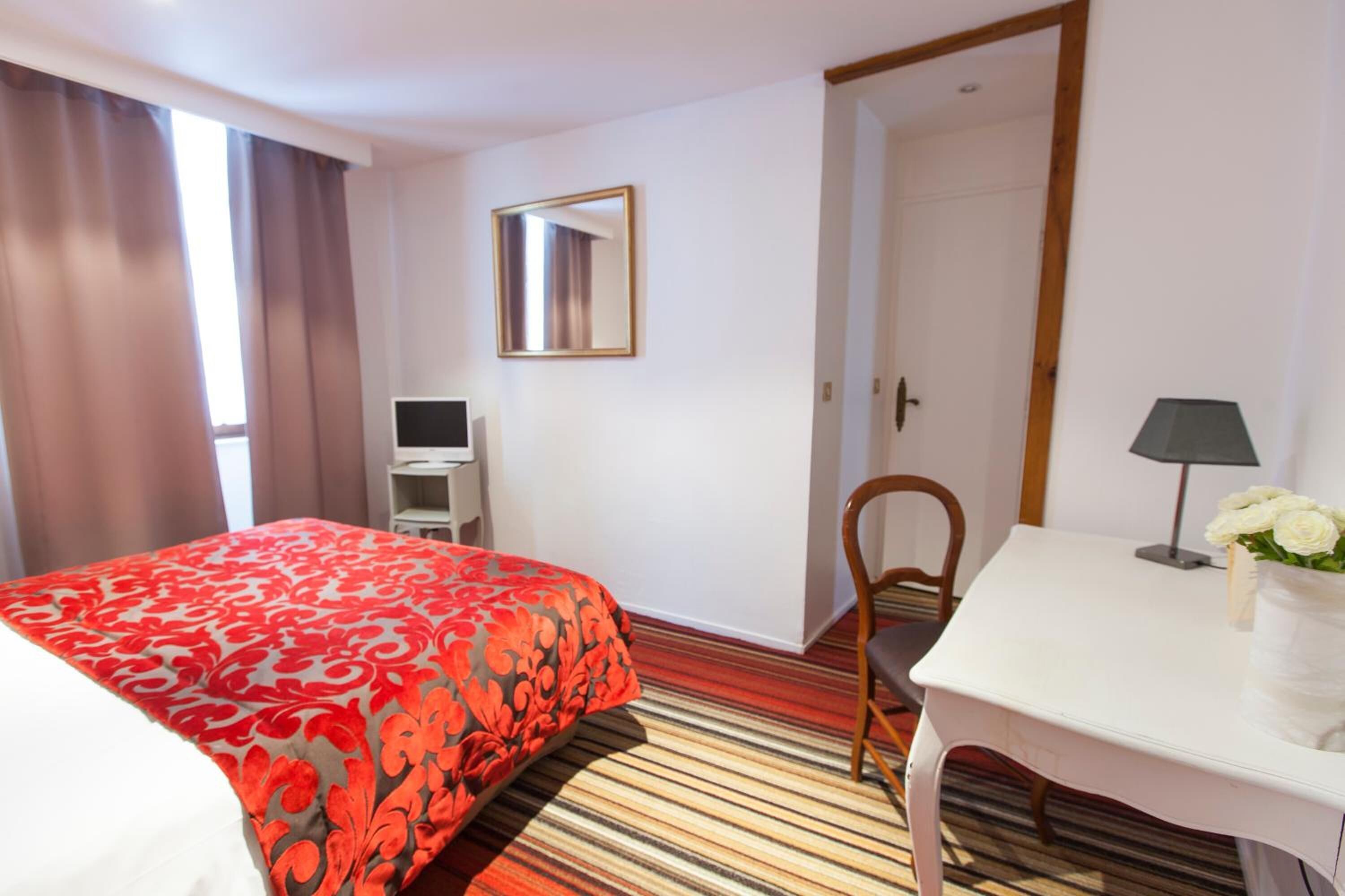 A hotel room with a bed, a desk, a chair, a mirror, and a door.