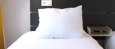 Un lit bien fait, avec un oreiller blanc et un magazine posé dessus. On y trouve une table de chevet avec une lampe et une prise électrique.
