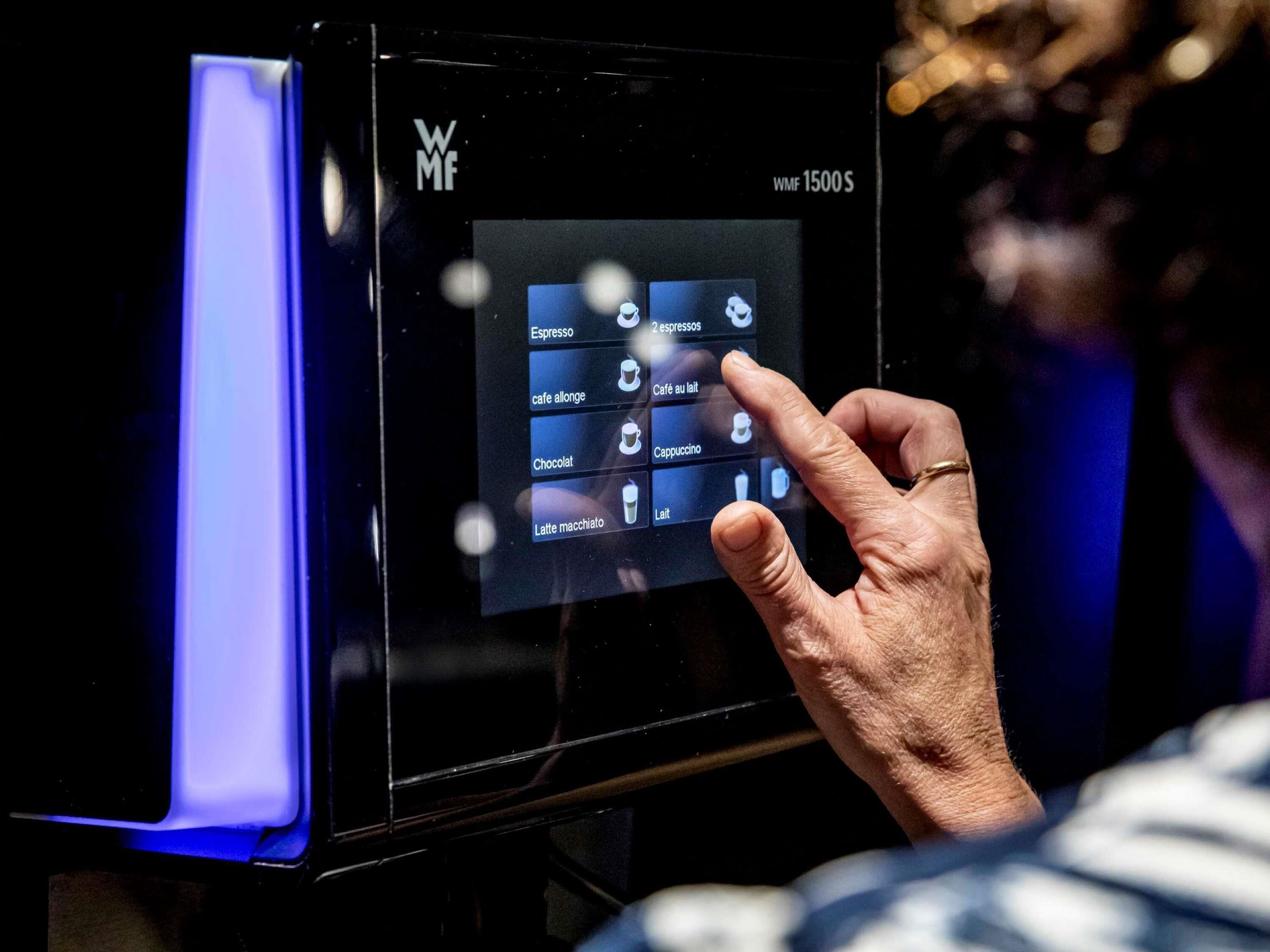 Una persona interactuando con una pantalla táctil en un electrodoméstico negro, posiblemente una cafetera, con opciones para espresso, café au lait, capuchino y latte macchiato.