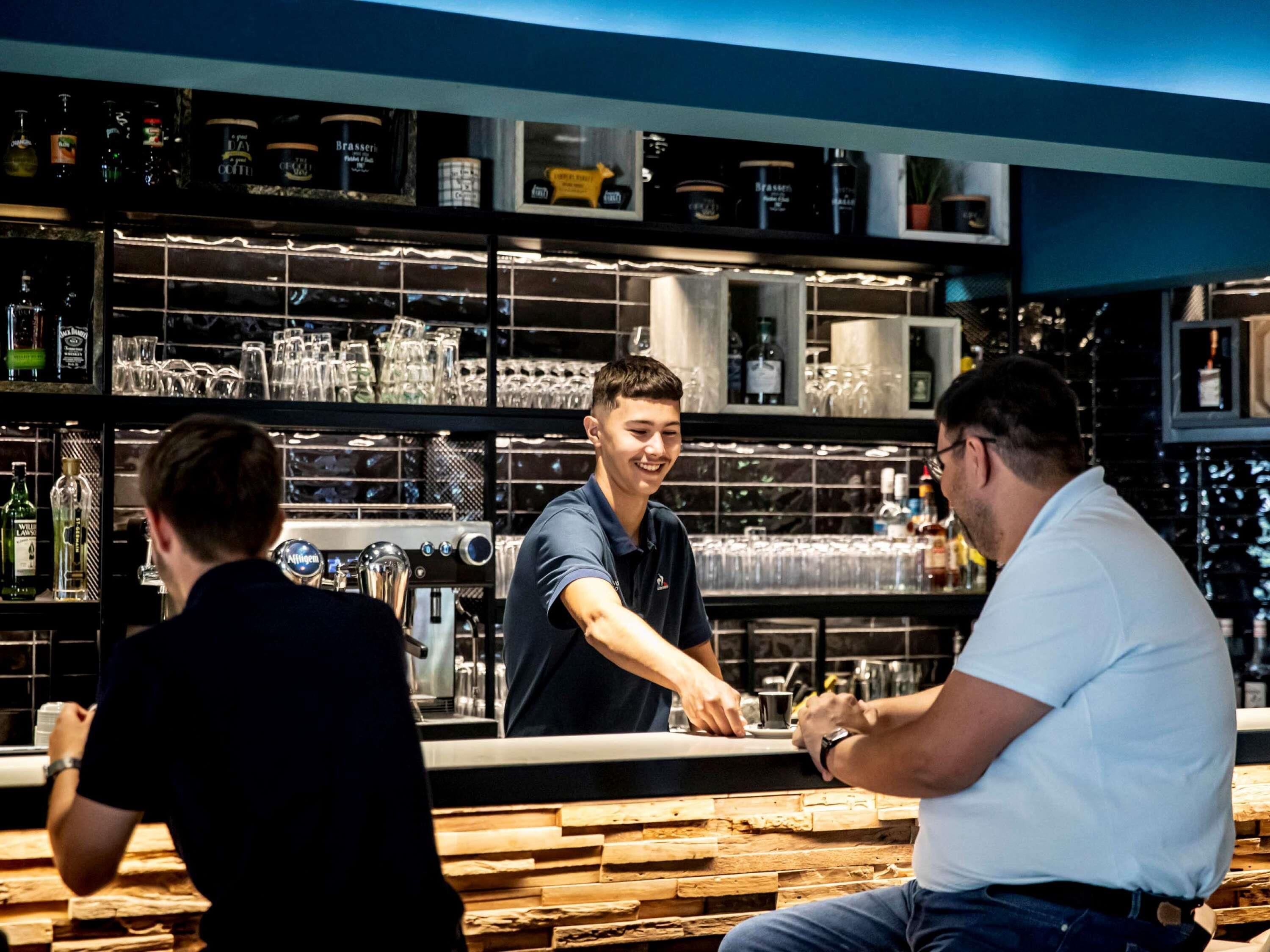 Zona de bar con estantes llenos de botellas y vasos, barra y una persona sirviendo bebidas.