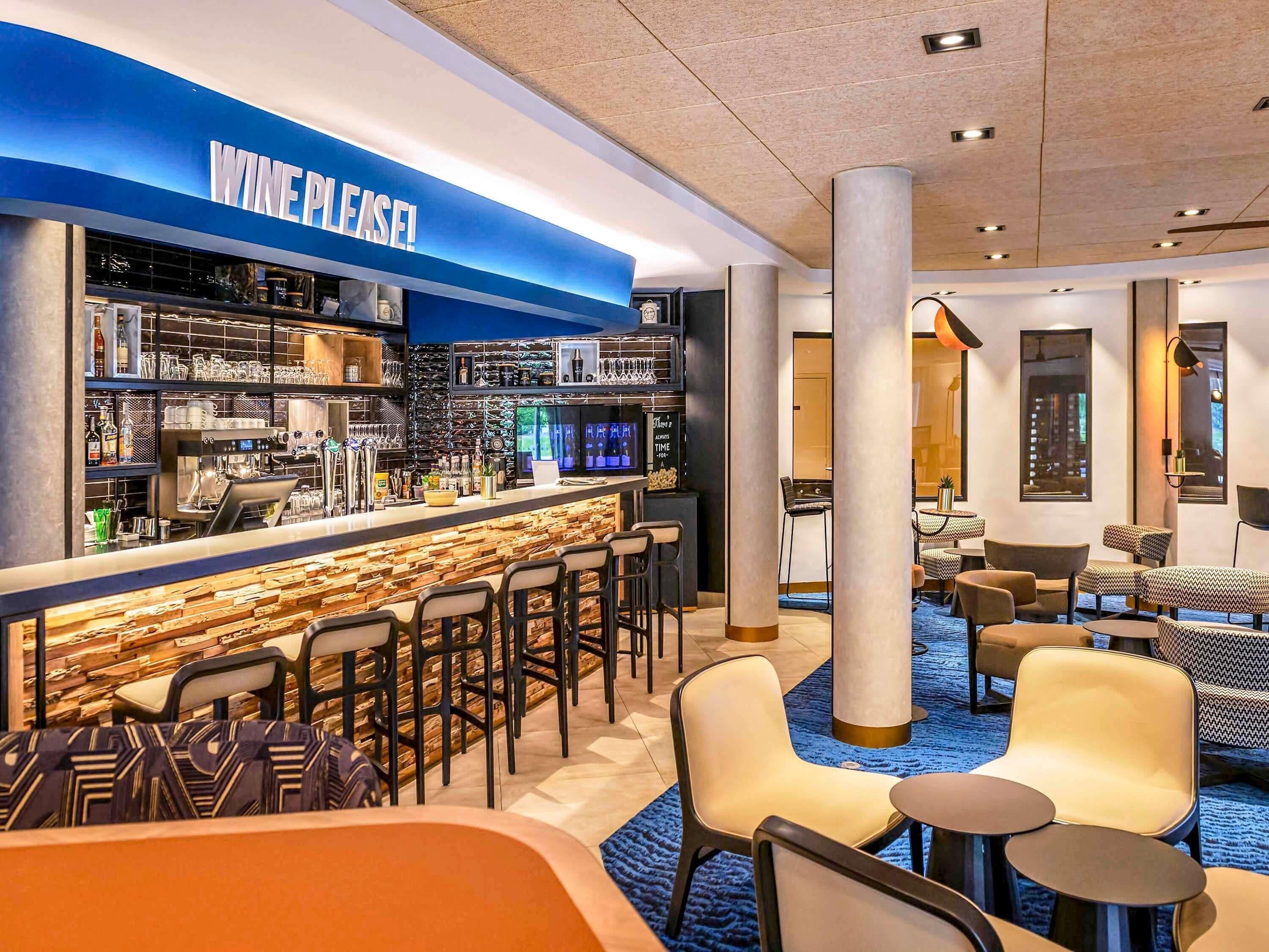 A bar area with a 'WINE PLEASE!' sign, bar stools, and a seating area with round tables and chairs.
