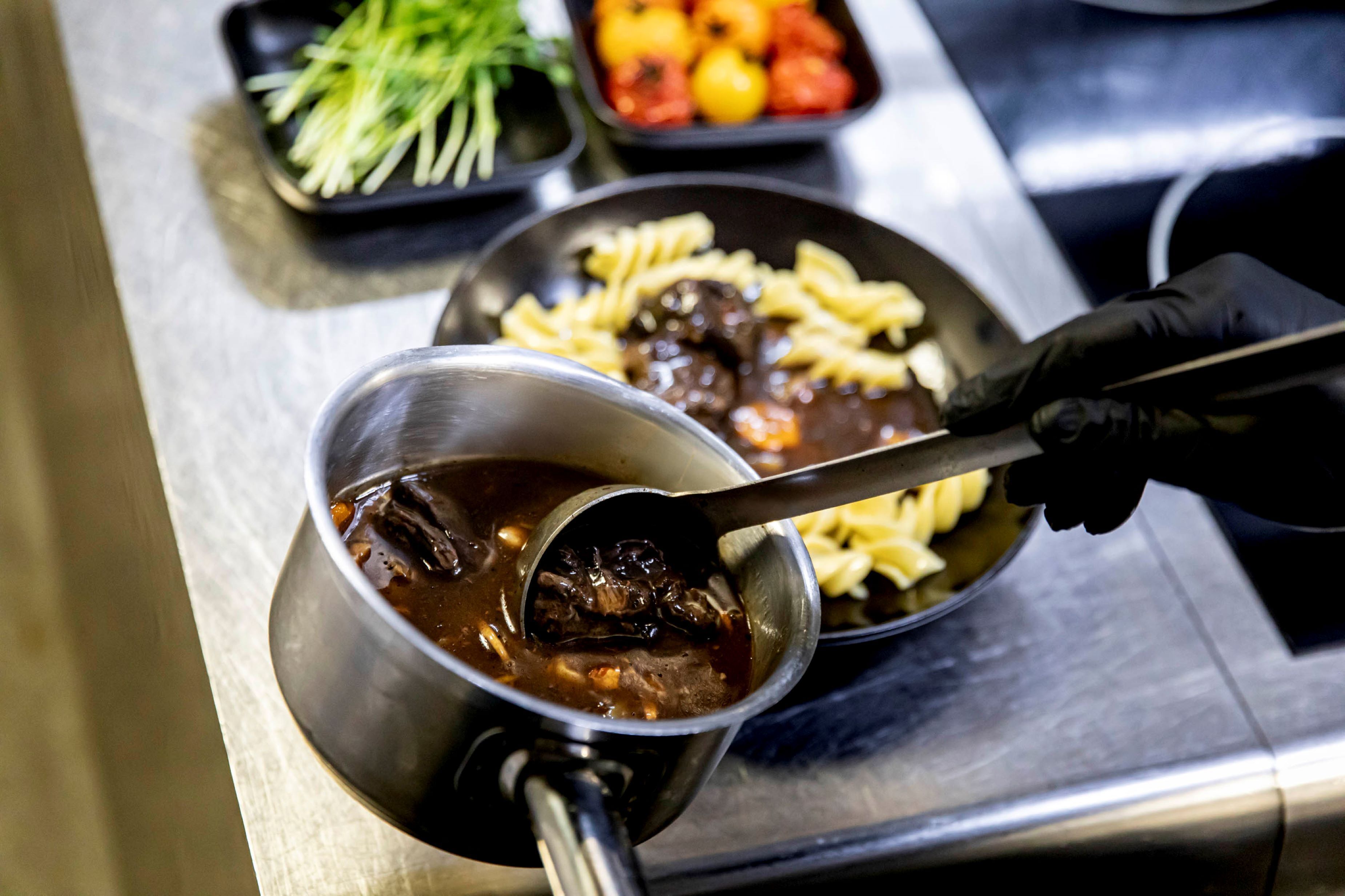 Eine Person schöpft eine dunkle Suppe aus einem Metalltopf, im Hintergrund sind Pasta und Gemüse zu sehen.
