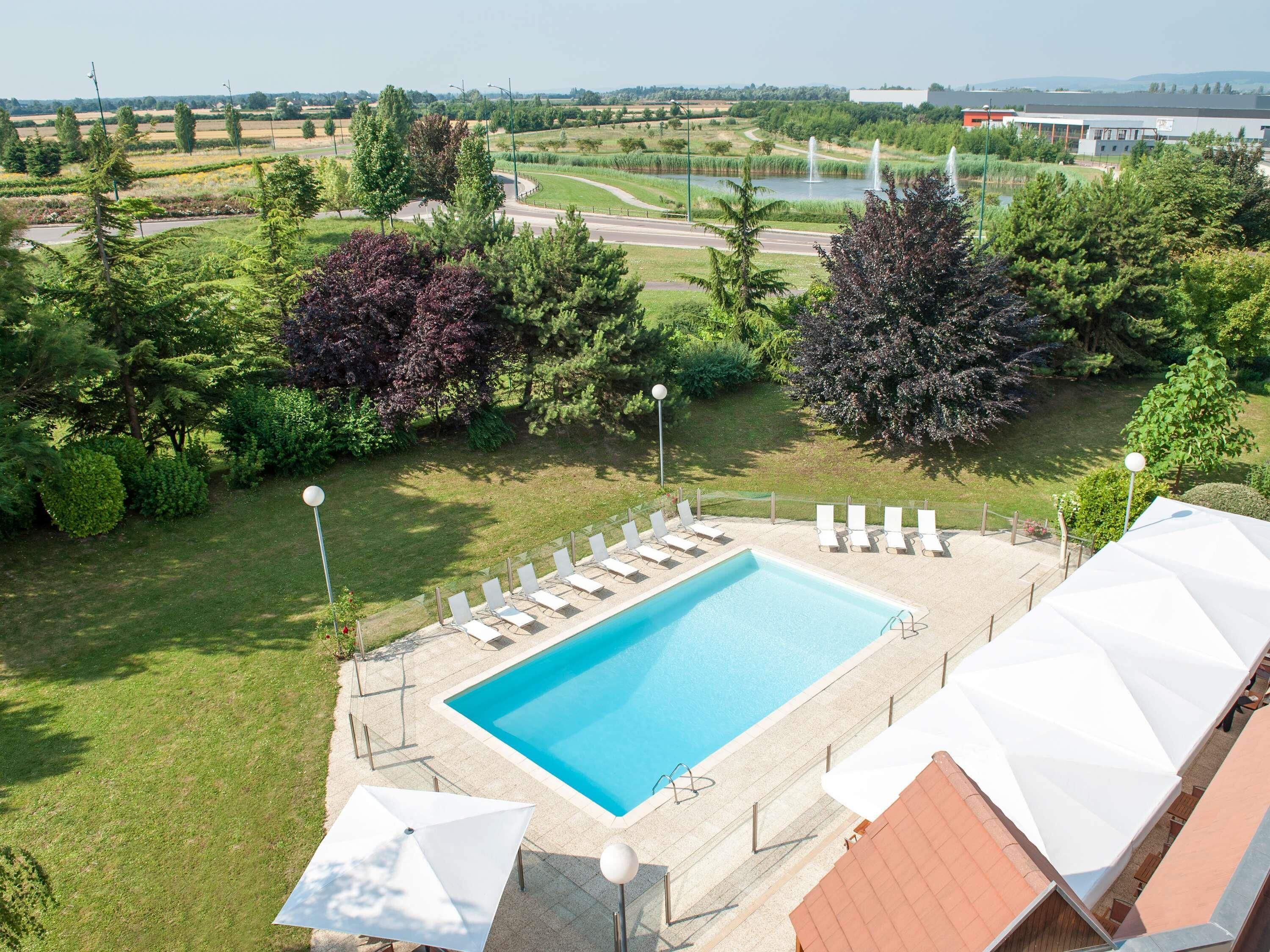 A swimming pool with lounge chairs, surrounded by greenery and a view of a park with fountains.