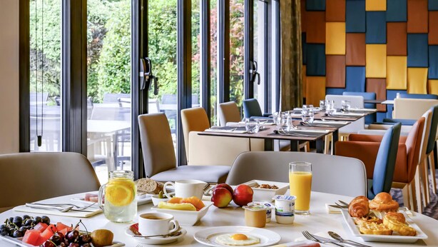 Una escena de desayuno con una mesa dispuesta para dos personas, incluyendo platos de fruta, huevos y pasteles, y vistas al exterior a través de amplias ventanas.
