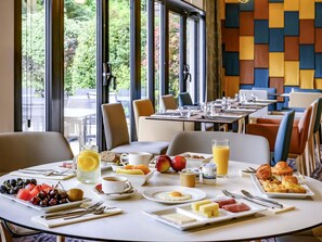 Una escena de desayuno con una mesa dispuesta para dos personas, incluyendo platos de fruta, huevos y pasteles, y vistas al exterior a través de amplias ventanas.