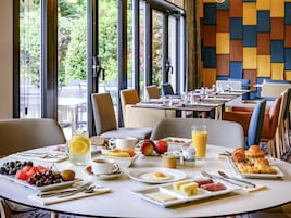 Una escena de desayuno con una mesa dispuesta para dos personas, incluyendo platos de fruta, huevos y pasteles, y vistas al exterior a través de amplias ventanas.