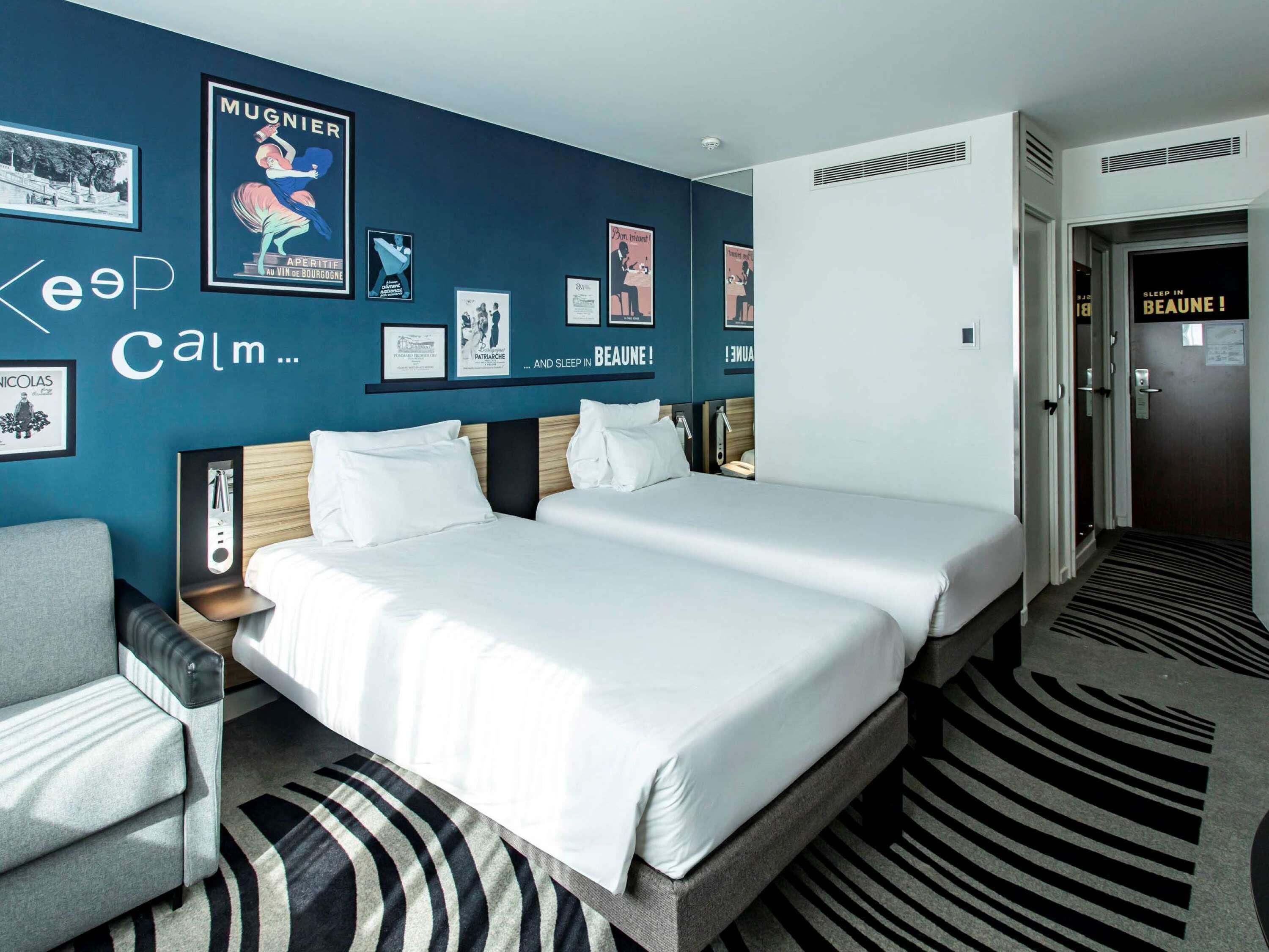 Habitación de hotel con dos camas, una silla, un escritorio y una pared decorada con cuadros y carteles.