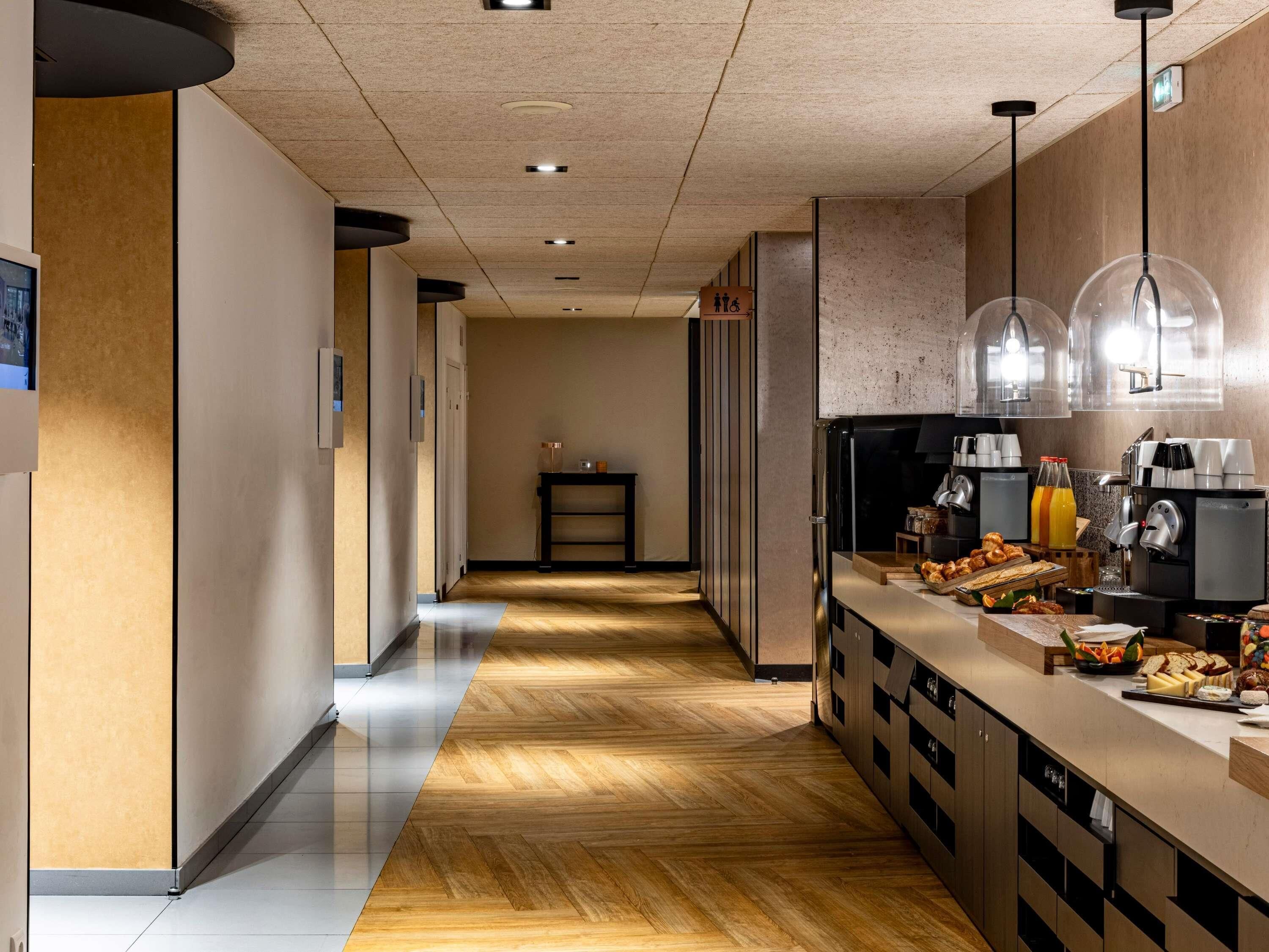Corridoio d'albergo con pavimento in legno, angolo cottura con macchine da caffè e un piccolo tavolo con una bottiglia.