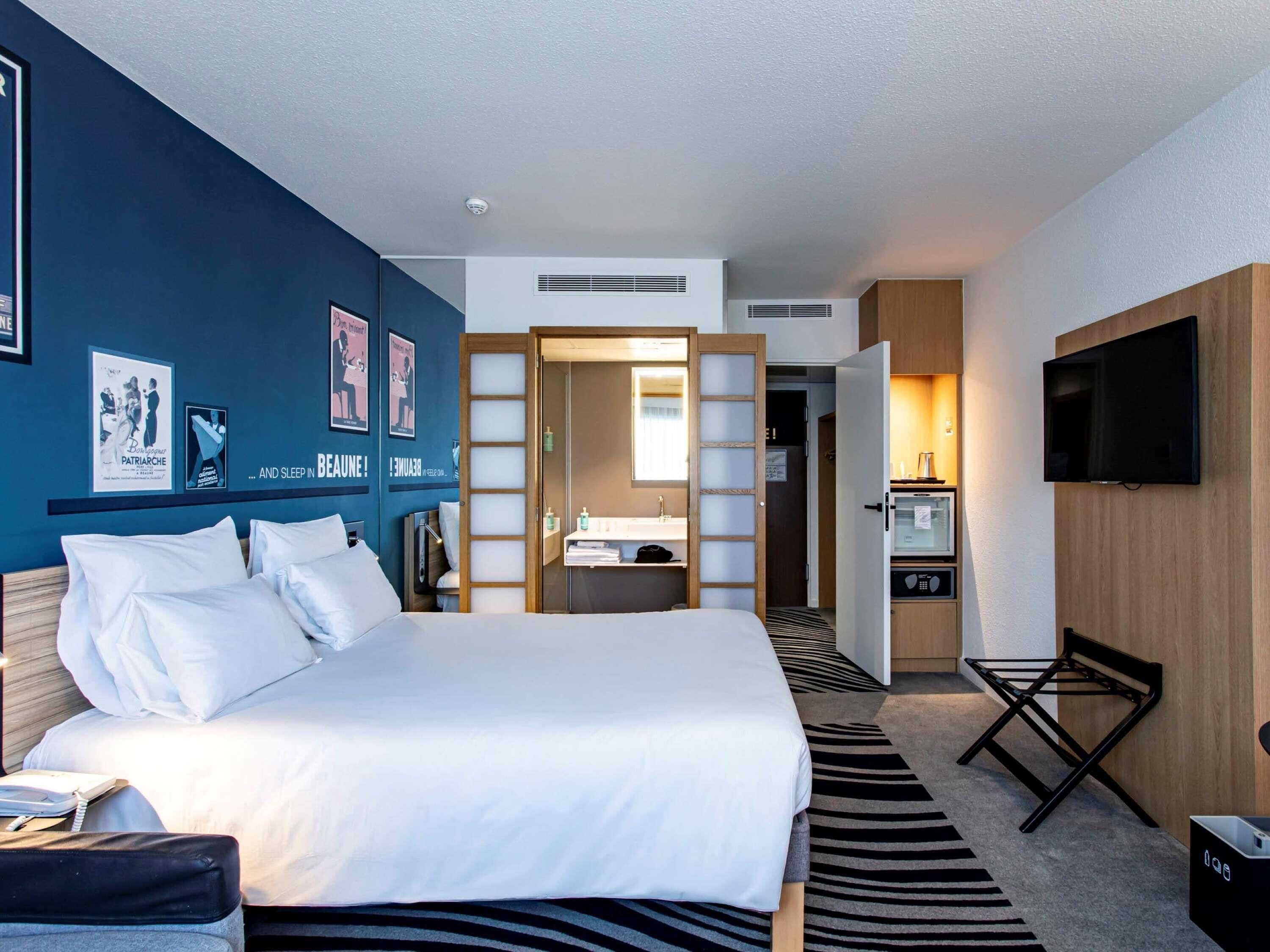 A hotel room with a bed, a desk, a TV, and a bathroom visible through an open door.