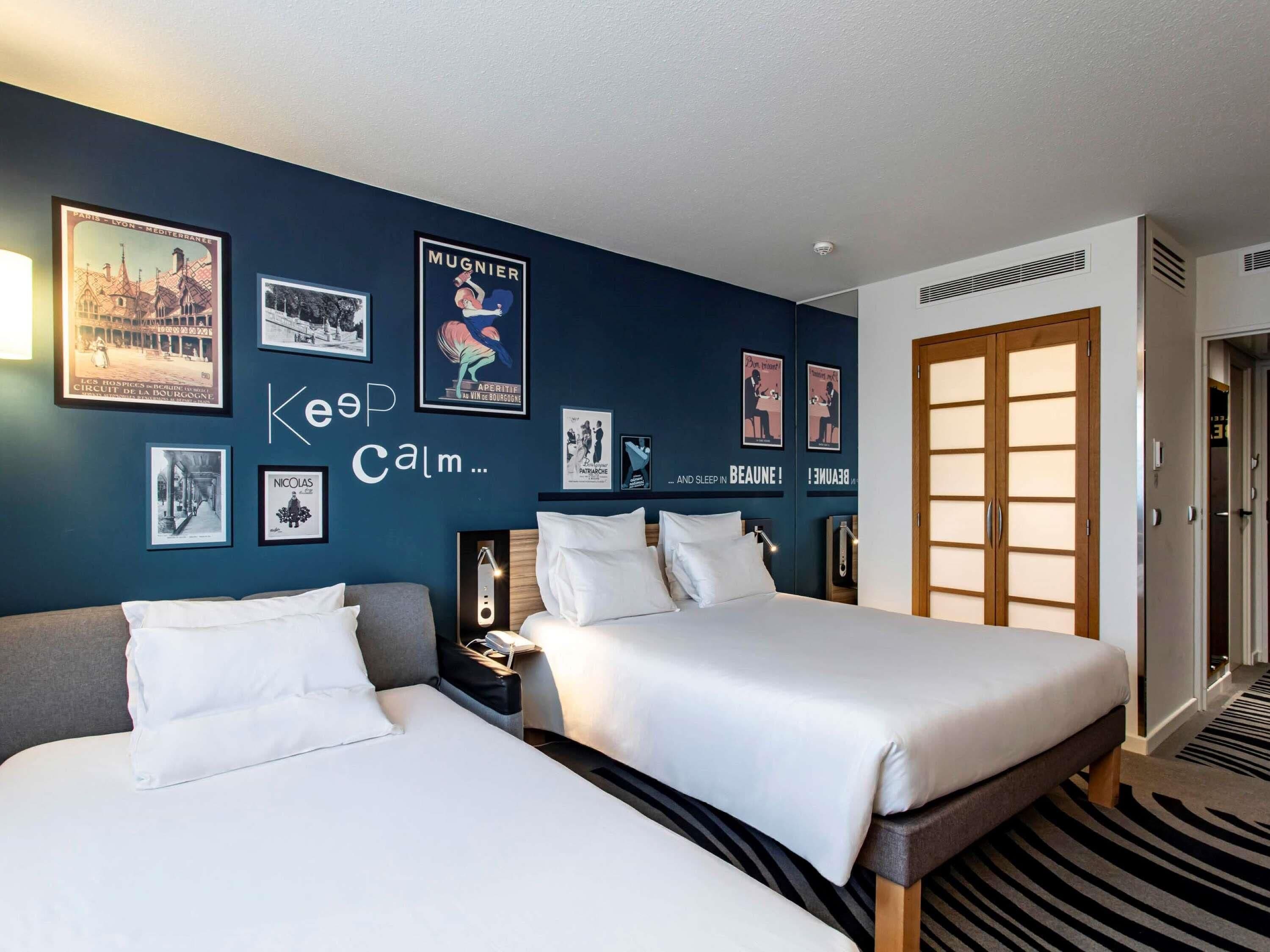 A hotel room with two beds, a dark blue wall with framed pictures, a wooden door, and a patterned carpet.