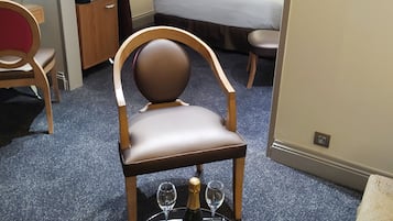 Una habitación de hotel con una cama, una mesita de noche con una botella y vasos, una silla y una pequeña mesa con una botella y vasos.