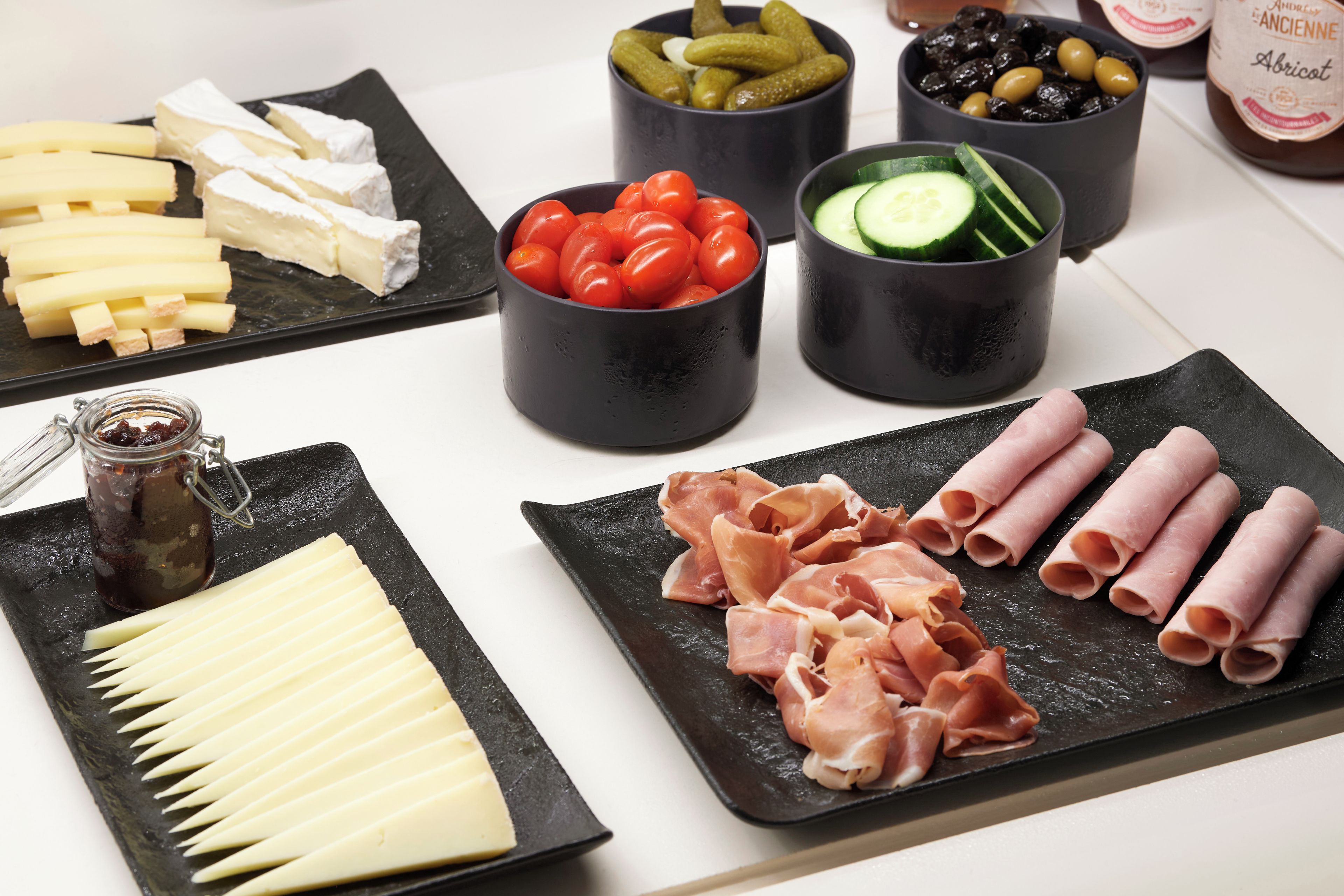 Diversos alimentos como queso, tomates, pepinos, aceitunas y embutidos dispuestos en platos y cuencos negros.