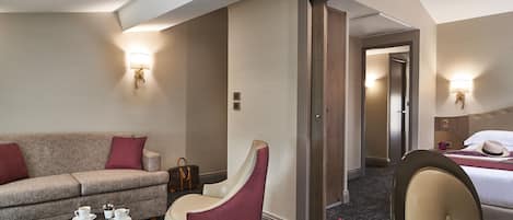 A hotel room with a sofa, armchair, and a bed. There is a table with a cup and saucer, and a handbag on the floor.