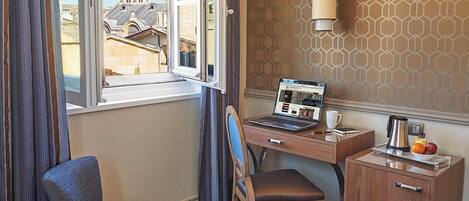 Une chambre d’hôtel avec un lit, un bureau avec un ordinateur portable, une chaise, une petite table avec une cafetière et des fruits, et une fenêtre avec des rideaux.