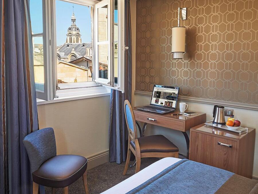 Habitación de hotel con cama, escritorio con computadora portátil, silla, pequeña mesa con cafetera y fruta, y ventana con cortinas.
