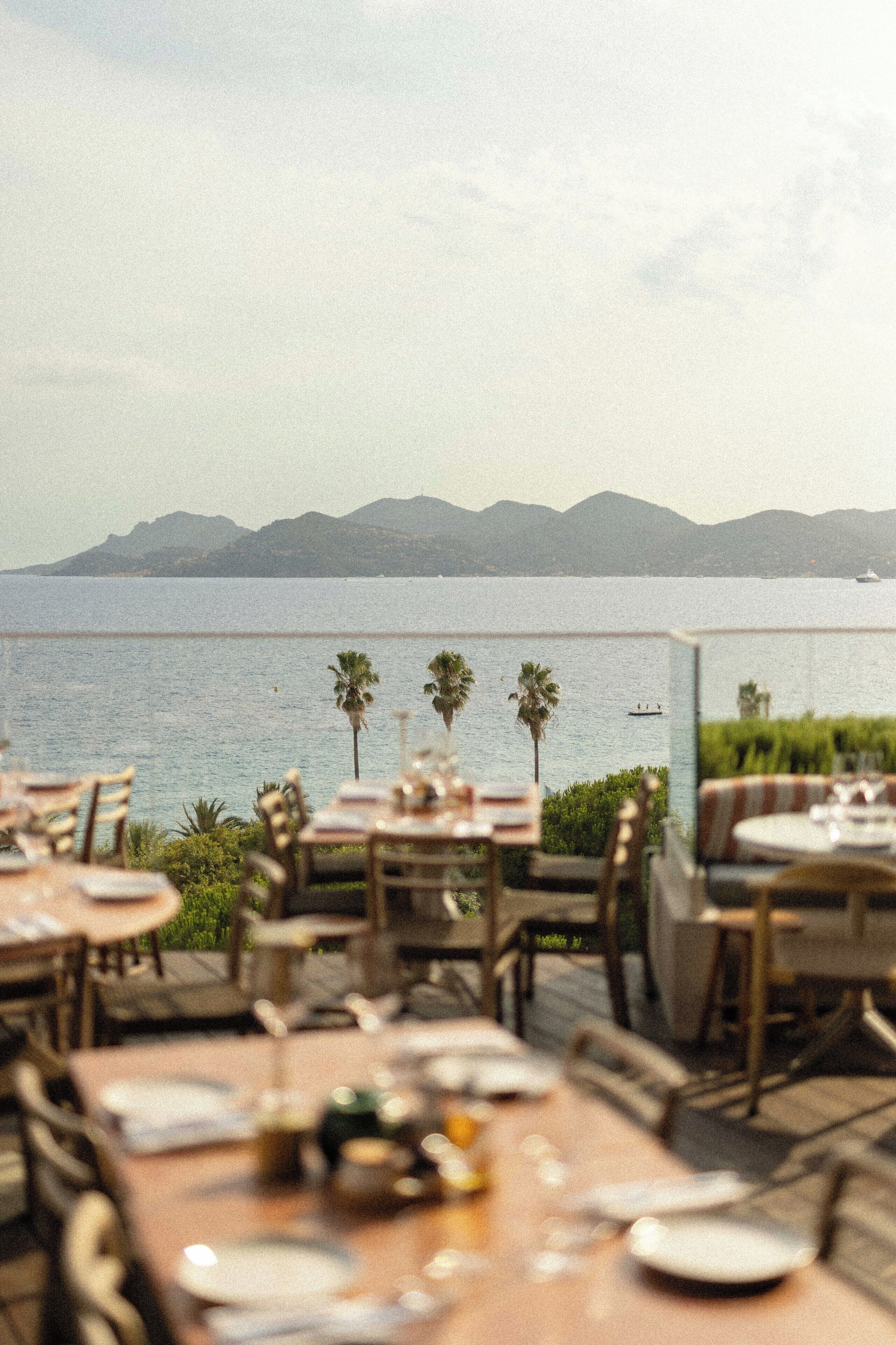 An outdoor dining area with tables and chairs, overlooking a body of water and distant mountains.