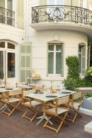 Een terras met houten tafels en stoelen, een witte balustrade en een groene haag.
