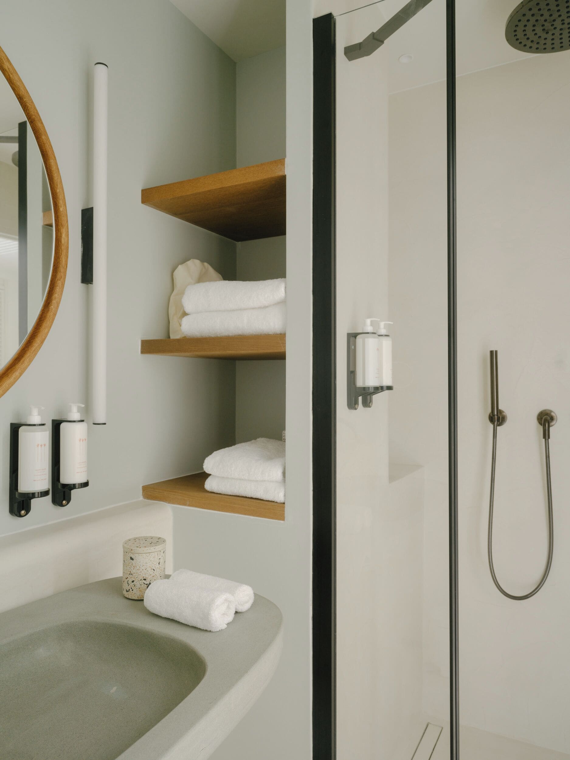 A modern bathroom with a glass-enclosed shower, a round mirror, and wooden shelves holding towels.