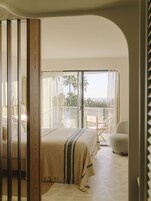 A bedroom with a large bed, a chair, and a balcony with a view of palm trees.