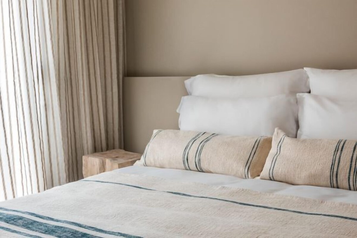 A bed with white bedding and striped pillows, a wooden nightstand, and a striped curtain.