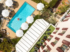 Luchtbeeld van een hotel met een zwembad, omringd door ligbedden en parasols, een dakterras en een gebouw met rode luiken.