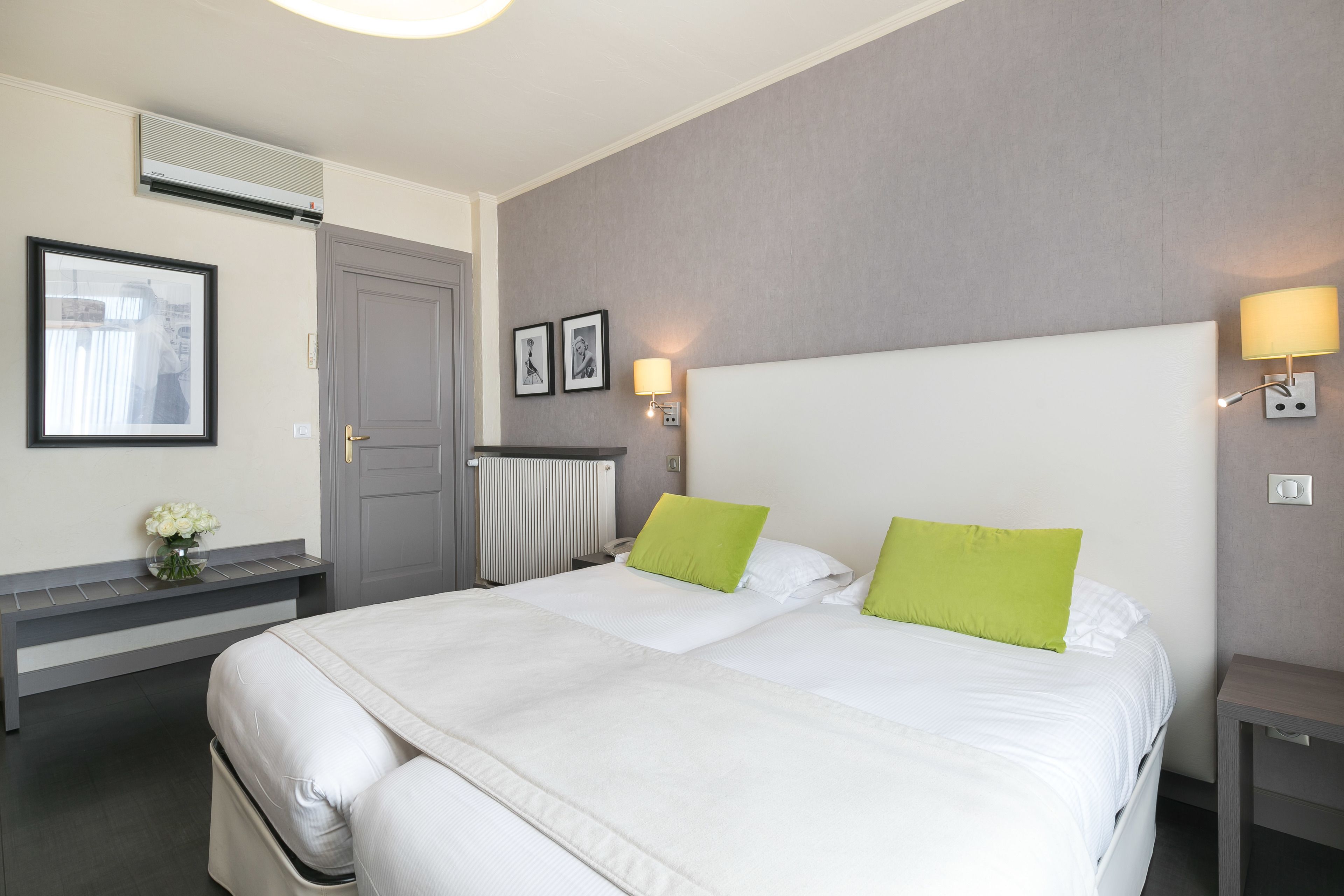 A hotel room with a large bed, a bedside table, a nightstand, a wall-mounted air conditioner, a framed picture, and a small vase with flowers.