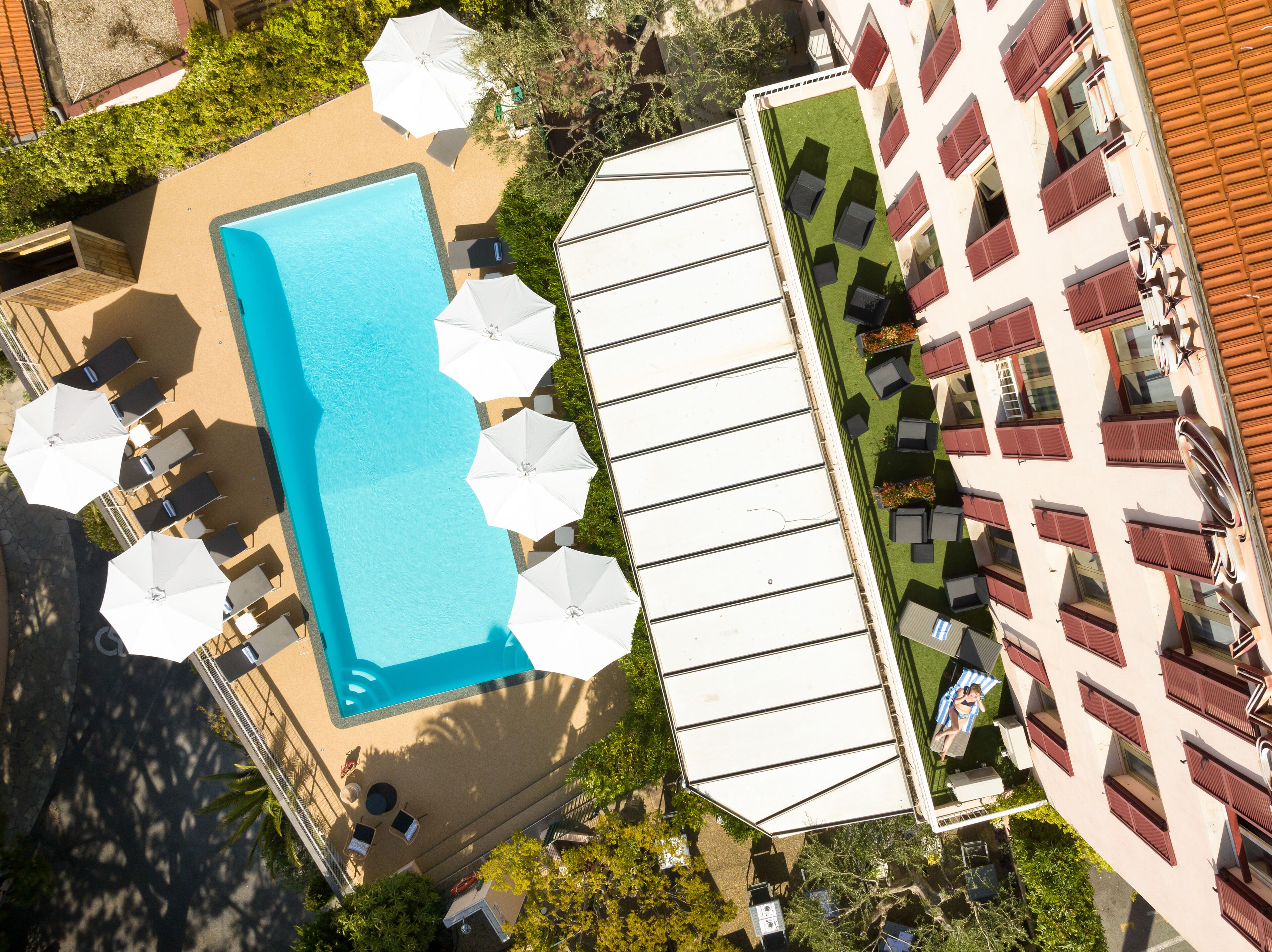 An aerial view of a hotel with a swimming pool surrounded by sun loungers and umbrellas, a rooftop bar with outdoor seating, and a building with red shutters.