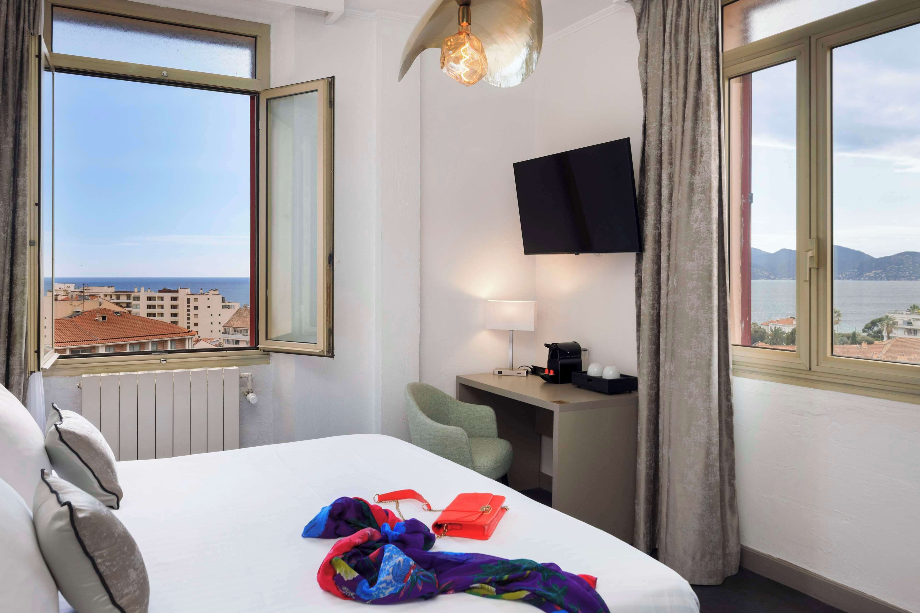 A hotel room with a bed, a desk, a chair, a TV, and a view of the sea and buildings.